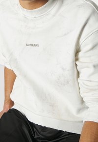 Bílá mikina s texturovaným vzorem, s příjemným ribovým výstřihem a manžetami. Na přední straně je umístěn kovový akcent s logem.