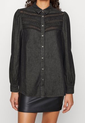 Femme portant un chemisier sombre à boutons avec un détail en dentelle sur le haut de la poitrine et des manches longues bouffantes, associé à une mini-jupe en cuir noire.