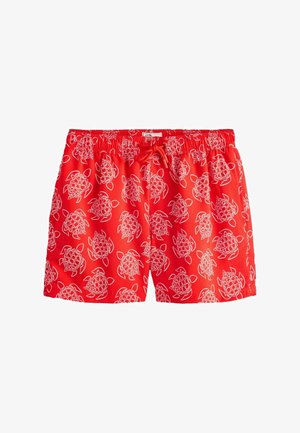 Gros plan sur une personne portant un short de bain rouge vif à motif de tortue blanche et à taille élastique avec cordon de serrage.