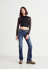Zwart cropped langemouwen top met mesh mouwen, gecombineerd met blauwe flared jeans. Het model draagt bruine klompen, wat een casual outfit toont.