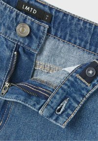 Jeansowe szorty w średnim niebieskim kolorze z zamkiem błyskawicznym, zapięciem na guzik i ciemnymi szwami. Zawierają małe elementy dekoracyjne oraz postrzępione krawędzie.