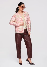 Femme portant une veste fleurie rose et beige, un top crème, un pantalon en cuir marron foncé, des talons hauts, de grandes lunettes de soleil et des boucles d’oreilles remarquables.