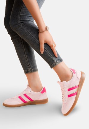 Personne portant un jean sombre et des baskets roses avec des rayures rose vif, pliant une jambe et touchant le mollet avec la main.