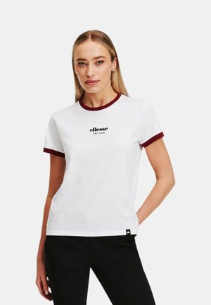 Donna che indossa una maglietta bianca con rifiniture rosso scuro su maniche e collo, con la scritta "ellesse EST. 1959" sul petto, abbinata a pantaloni neri.