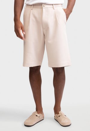 Shorts beige mi-longueur avec plis portés par une personne en chaussures beiges sans lacets, debout devant un fond clair uni.
