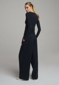 Granatowa bluzka z długim rękawem i szerokie, prążkowane spodnie. Luźny krój z gładką teksturą. Modelka stoi z rękami w kieszeniach, lekko odwrócona.