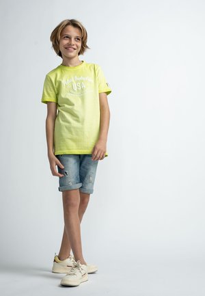 Lys grøn T-shirt med et hvidt logo, parret med slidte denimshorts. Casual hvide sko med røde og gule accenter.