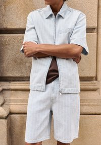 Personne portant une chemise à manches courtes bleu clair avec fermeture éclair et un short assorti, debout les bras croisés contre un mur en pierre texturée.