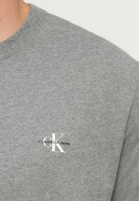 Szürke pamut póló rövid ujjakkal és kerek nyakkivágással. Kis fehér „CK” logóval és „Calvin Klein Jeans” felirattal a mellkason.