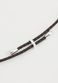Corda in pelle intrecciata marrone con accenti in metallo argentato; presenta una chiusura liscia e cilindrica e una perla decorativa arrotondata.