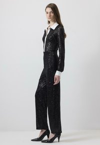 Svart paljett jumpsuit med lång ärm, krage i vitt och rak passform, matchad med svarta högklackade skor.
