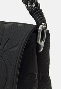 Schwarze Handtasche aus strukturiertem Material mit geprägtem Blumenmuster und geflochtenem Riemen sowie silbernem Hardware-Akzent.