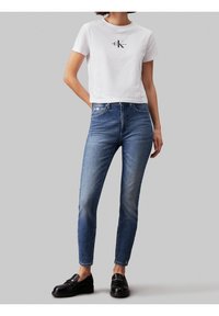 Vit croppad t-shirt med "CK Jeans"-logotyp, parat med åtsittande mörkblå denimjeans och svarta loafers, som visar rena linjer och en avslappnad stil.