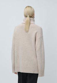 Μπεζ υπερμεγέθης πουλόβερ με ψηλό γιακά, ραμμένες μανσέτες και στρίφωμα. Μαλακό, υφασμένο ύφασμα με υφή. Εικόνα από το πίσω μέρος.