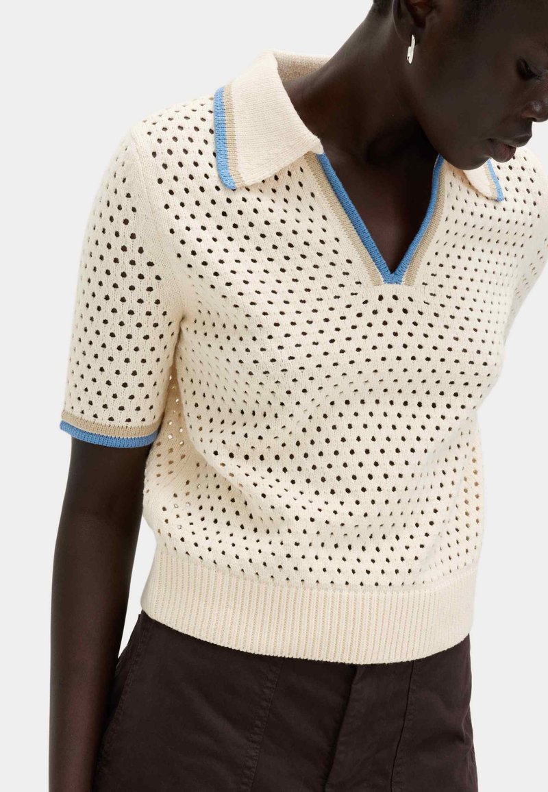 Persona de piel oscura que lleva un top de punto de manga corta color crema con ribete azul y beige y escote en V, combinado con pantalones oscuros.