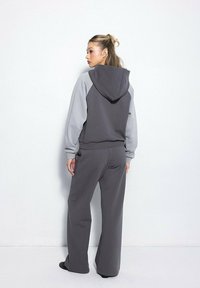 Kobieta stojąca tyłem, ubrana w dwukolorowy szary hoodie z jasnoszarymi rękawami oraz pasującymi szerokimi spodniami na białym tle.