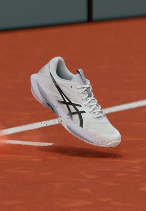 White athletic shoe with black logo mid-air above red clay tennis court, kicking up small clay particles near white court line.
