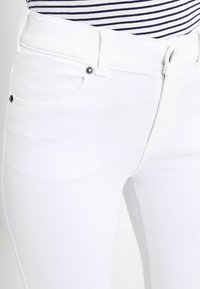 Calças de ganga brancas com uma textura suave, design de cinco bolsos e detalhes em metal. A cintura apresenta um fecho padrão com botão e fecho de correr.