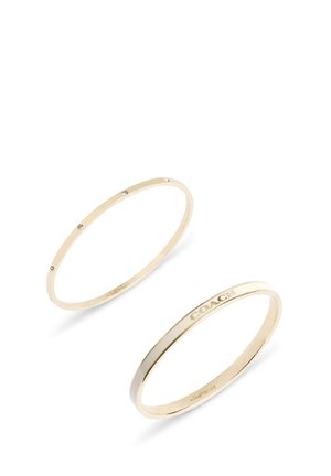 To slanke guldbangles, den ene med små indlejrede krystaller, den anden graveret med ordet "COACH" på en hvid baggrund.