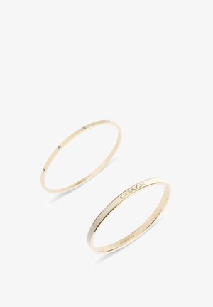 Due sottili braccialetti rigidi d'oro, uno con piccoli cristalli incastonati, l'altro inciso con la parola "COACH" su uno sfondo bianco.