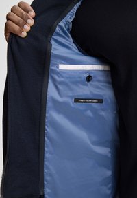 Wnętrze granatowej kurtki ma gładką, jasnoniebieską podszewkę, czarny guzik oraz kieszeń z białym akcentem. Etykieta głosi "NAJLEPSZA WŁOSKA TKANINA."