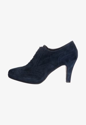 Chaussure à talon haut en daim bleu marine avec un bout fermé, des détails en brogue décoratifs et un talon fin. Texture lisse et forme élégante.