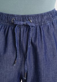 Joggers en denim avec une taille élastique, un cordon de serrage et des œillets en argent. Présente une couleur bleu moyen et un tissu doux et texturé.