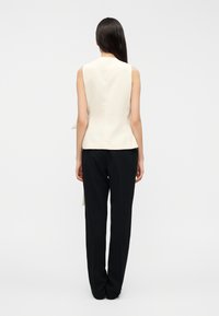 Αμάνικο κρέμ τοπ με εφαρμοστό σχέδιο, συνδυασμένο με μαύρο παντελόνι με ίσια γραμμή. Λείο υλικό, χωρίς ορατά μοτίβα ή λεπτομέρειες.