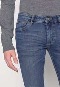 Tumman siniset farkut, joissa on klassinen viiden taskun muotoilu, suora leikkaus ja hienovaraiset ompeluyksityiskohdat saumoissa.