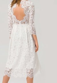 Bílé krajkové šaty s tříčtvrtečními rukávy, hlubokým výstřihem na zádech a lemováním s vlnkami. Mají složité květinové vzory po celé ploše.