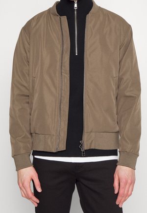 Veste bomber marron avec poignets et ourlet côtelés portée sur un pull noir zippé et un tee-shirt blanc, associée à un pantalon noir.
