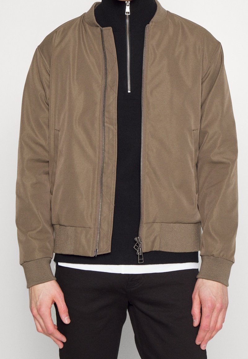 Veste bomber marron avec poignets et ourlet côtelés portée sur un pull noir zippé et un tee-shirt blanc, associée à un pantalon noir.