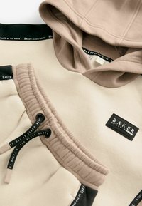 Sudadera crema con capucha color canela, cintura elástica y detalles en negro. Presenta una etiqueta con el logo y cordones para un ajuste ajustable.