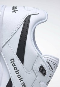White synthetic sneaker with black accents, textured surface, Reebok logo, padded collar, and contrasting stitching details.