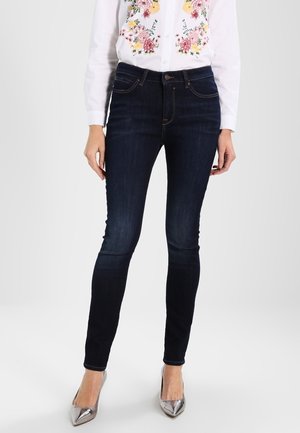 Kobieta ubrana w ciemne obcisłe dżinsy, białą koszulę z kwiatowym haftem oraz srebrne, szpiczaste wysokie obcasy, stojąca na tle jednolitego tła.