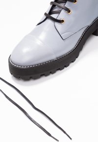 Botte en cuir gris clair avec semelle épaisse à crampons noire et lacets noirs, un lacet partiellement passé à travers des œillets dorés, sur fond blanc.