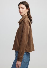 Bruine suède overhemdjack met een getextureerde zak, knoopsluiting en een kraag. Draagt met lichtblauwe denim jeans.