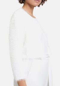 Biały, krótki kardigan o puszystej fakturze, z długimi rękawami zwieńczonymi frędzlami, w połączeniu z prostą białą odzieżą pod spód.