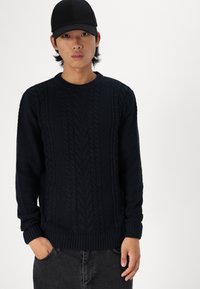 Suéter de punto azul marino oscuro con un patrón de trenzas, cuello redondo y puños acanalados. Combinado con pantalones gris oscuro, con las manos en los bolsillos.