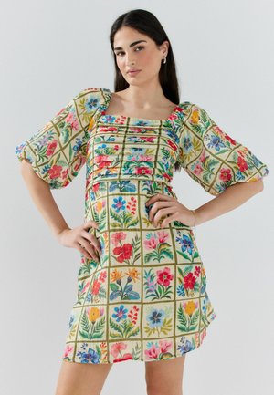 Kobieta z długimi ciemnymi włosami, ubraną w krótką beżową sukienkę z kolorowymi kwadratowymi wzorami kwiatowymi i bufkami na półdługich rękawach, pozuje z rękami na biodrach.