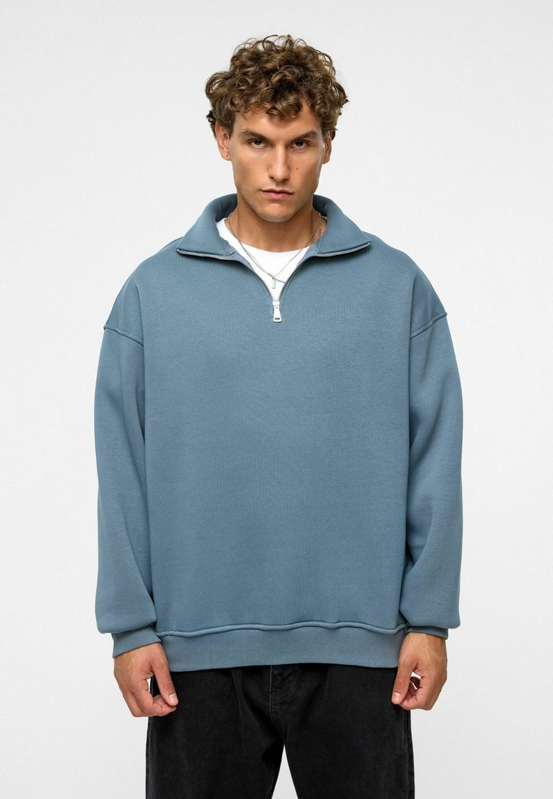 Lichtblauwe fleece pullover met een ritskraag, verlaagde schouders, geribbelde manchetten en een relaxte fit. Gecombineerd met een wit t-shirt.