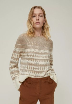 Maglione a maglia beige e crema con motivi geometrici e accenti metallici. Collo e polsini a coste. Modello che indossa pantaloni in velluto a coste marroni.