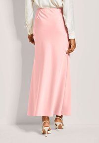 Lange, roze satijnen rok met een gladde textuur, voorzien van een aansluitende tailleband en een vloeiende, volle silhoeët. Draagt met witte sandalen met bandjes.