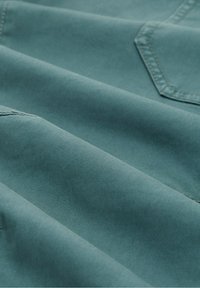 Tissu plié bleu sarcelle avec coutures visibles et vue partielle du bord d'une poche cousue en haut à droite.