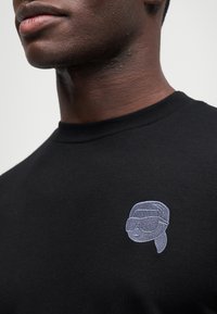 Μαύρο βαμβακερό μπλουζάκι με λαιμόκοψη, διαθέτει ένα ανοιχτό μπλε κεντητό σχέδιο προσώπου με γυαλιά ηλίου στο αριστερό στήθος.