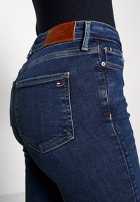 Los jeans de mezclilla azul oscuro presentan un parche de cuero marrón etiquetado con "TOMMY HILFIGER" en la cintura, con los bolsillos traseros mostrando un pequeño detalle del logo.