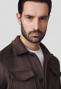 Brązowa wełniana kurtka z kołnierzem, dwoma przednimi kieszeniami i zapięciem na guziki. Nosi się ją na białym T-shircie, który ma gładką fakturę i dopasowany krój.