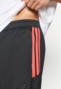 Μαύρο αθλητικό σορτς με κόκκινες ραβδώσεις, με τσεπάκι με φερμουάρ και ανάγλυφη μαύρη πιν ελιά στο κάτω μέρος.