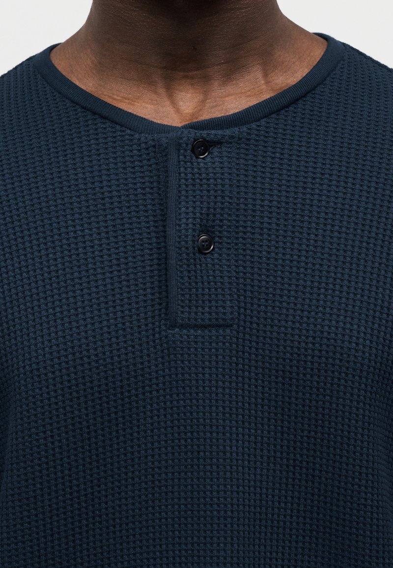 Pull-over bleu marine en maille gaufrée avec une surface texturée, doté d'un col rond et de deux boutons noirs à l'avant.