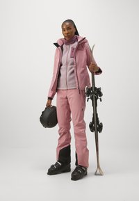 Tenue de ski rose composée d'une veste imperméable, de pantalons assortis avec des accents noirs et d'une couche douce en polaire. Elle tient des skis et un casque.